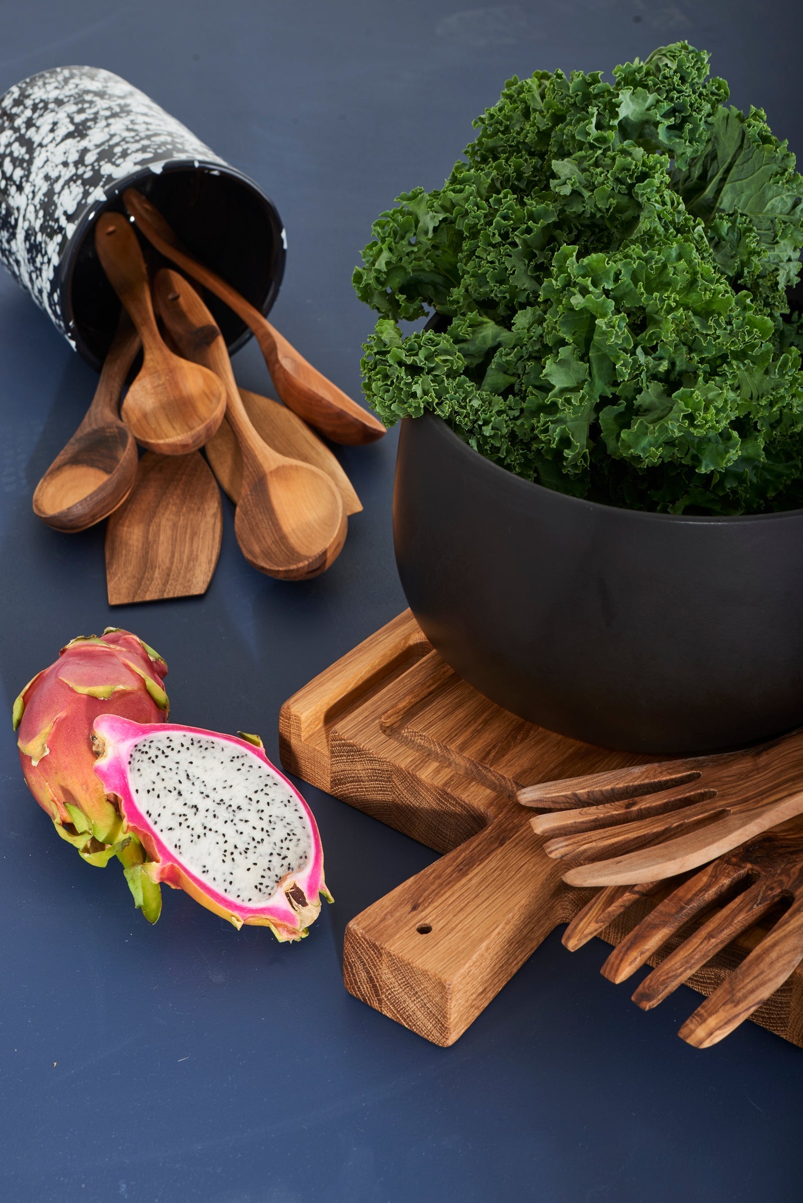 Some objects from around the kitchen are laid out on a dark blue background, including a black bowl filled with kale, wooden spoons and salad scoopers, a wood cutting board and a dragon fruit cut in half.